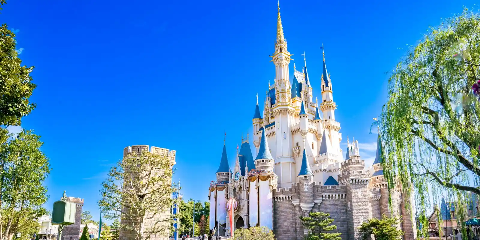 White and gold Cinderella Castle with blue-roofed turrets under a clear blue sky, framed by a stone tower on the left and a weeping willow on the right