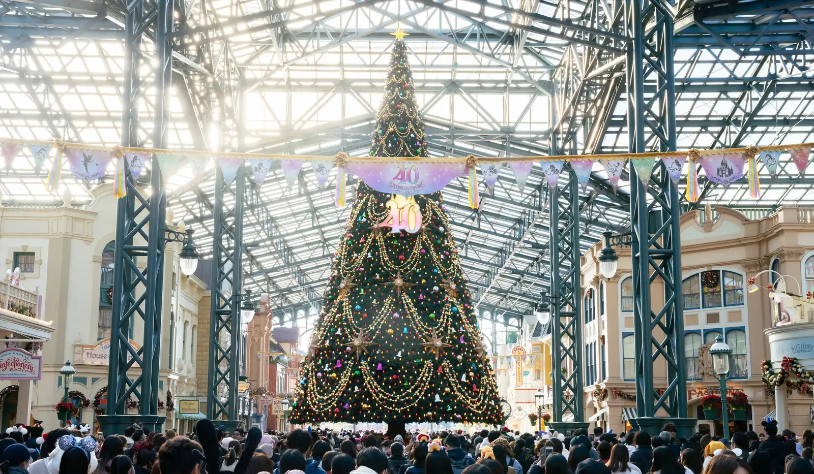 Glass-roofed shopping street with cream Victorian-style shopfronts and a tall decorated Christmas tree in the middle, crowds walking towards it