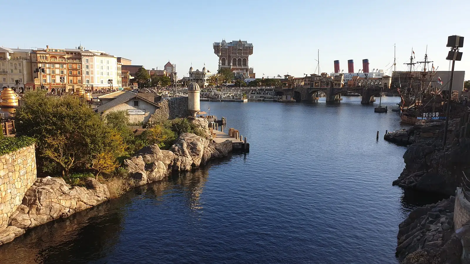 A calm channel of water lined with rocks and trees leading towards American Waterfront, where the tall Tower of Terror hotel building and the red funnels of a large ocean liner stand on the skyline