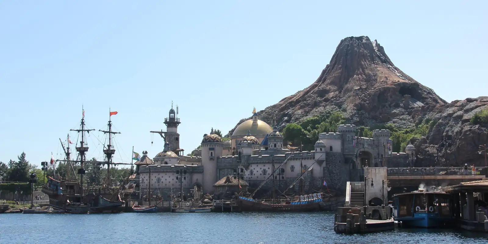 Mediterranean Harbor at Tokyo DisneySea: tall sailing ships and a domed stone fortress on the water, with the rocky volcano Mount Prometheus rising behind under a clear blue sky