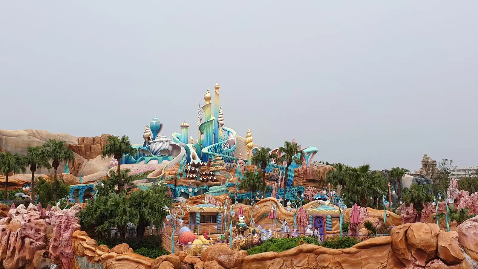 The colourful coral-and-shell spires of King Triton's castle at Mermaid Lagoon, with palm trees and pink rock formations in front under a grey sky
