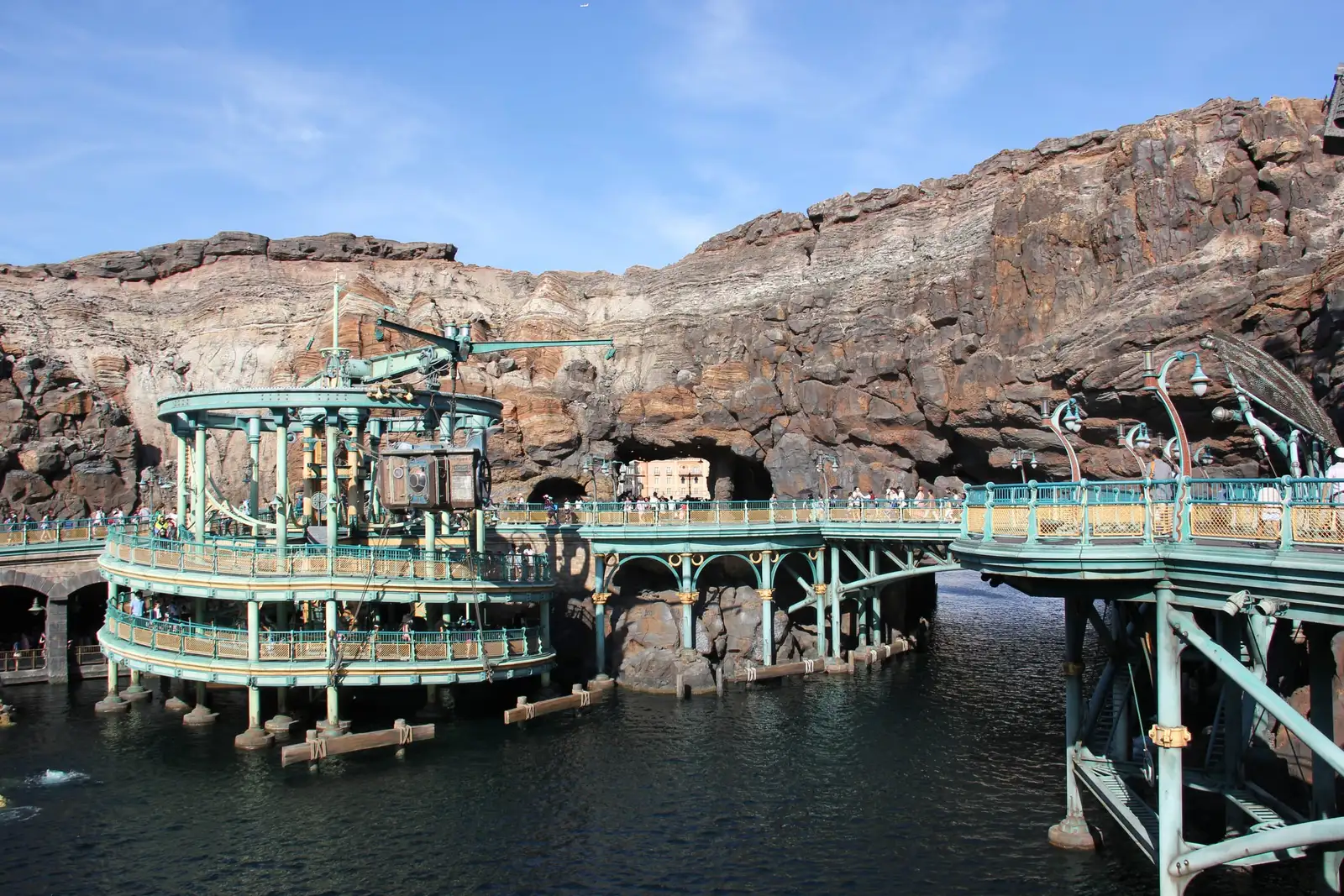 Teal iron walkways and a platform on stilts over dark water in Mysterious Island, surrounded by tall brown volcanic rock walls under a blue sky