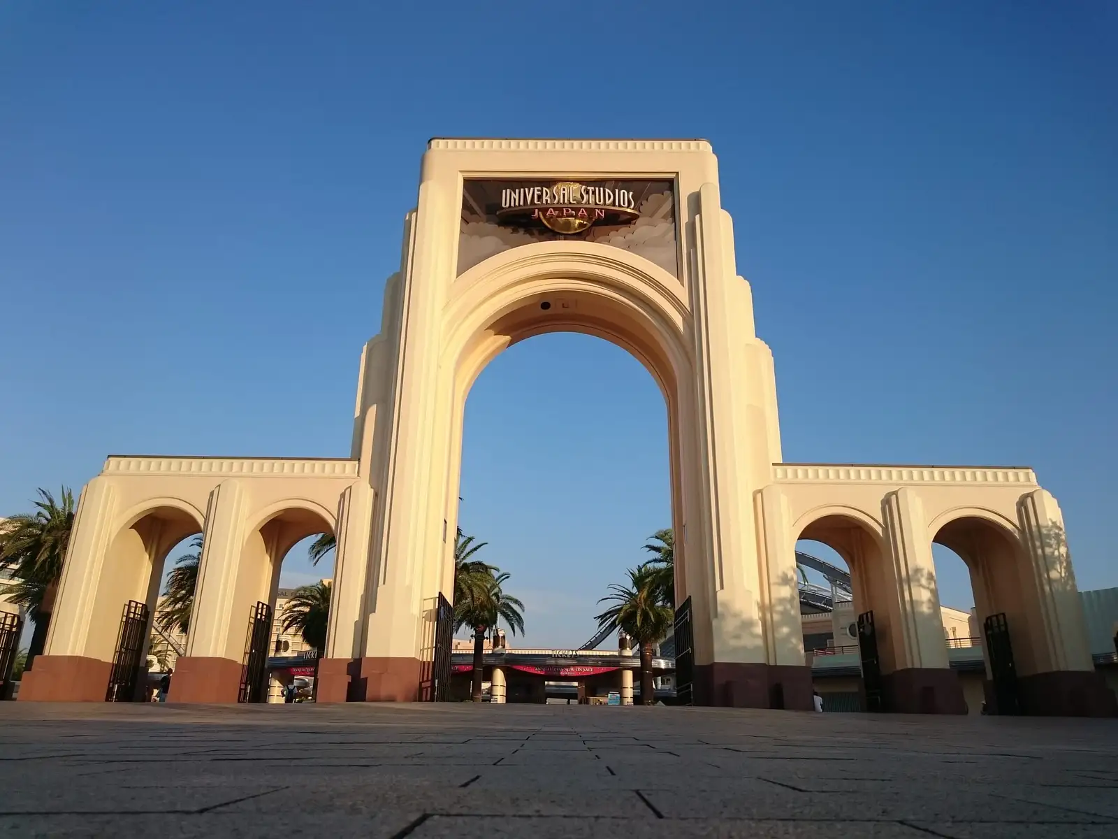 Кремовая тройная арка главного входа Universal Studios Japan под ясным голубым небом, за воротами — пальмы