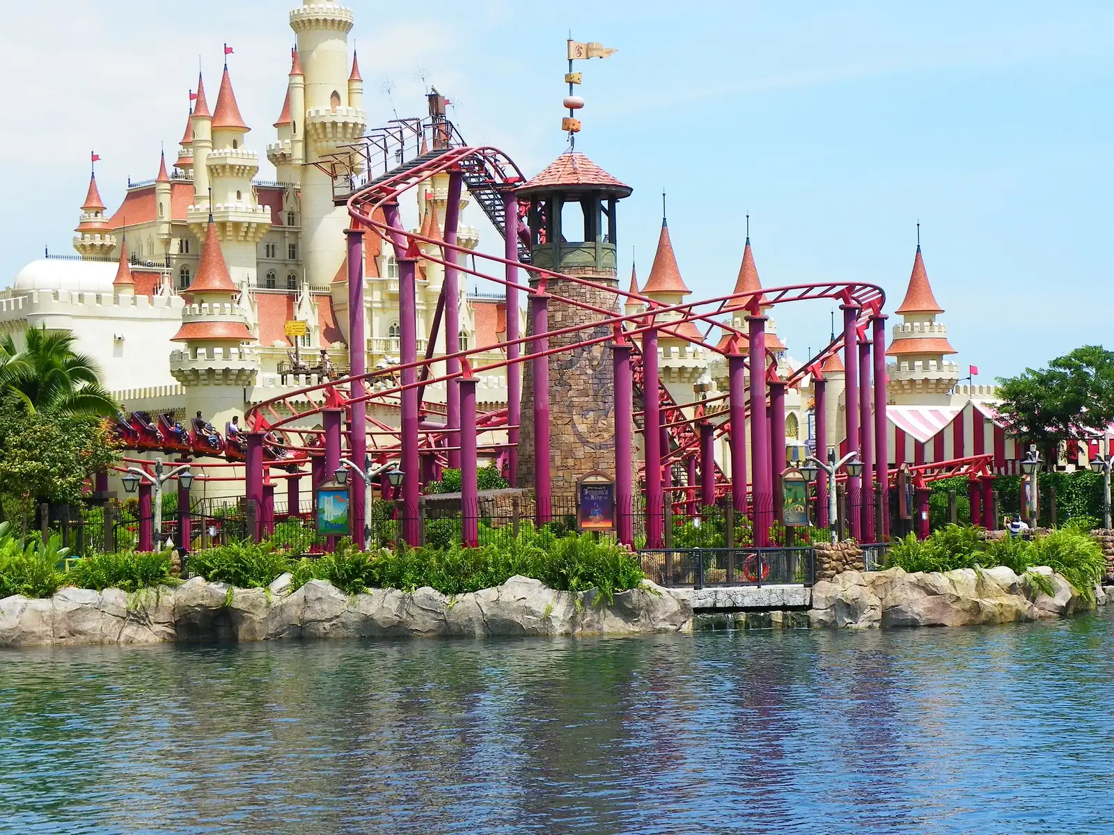 A cream-coloured fairy-tale castle with orange-red conical roofs behind a magenta-painted junior roller coaster that loops around a stone tower, with ferns, rocks and the lagoon in the foreground