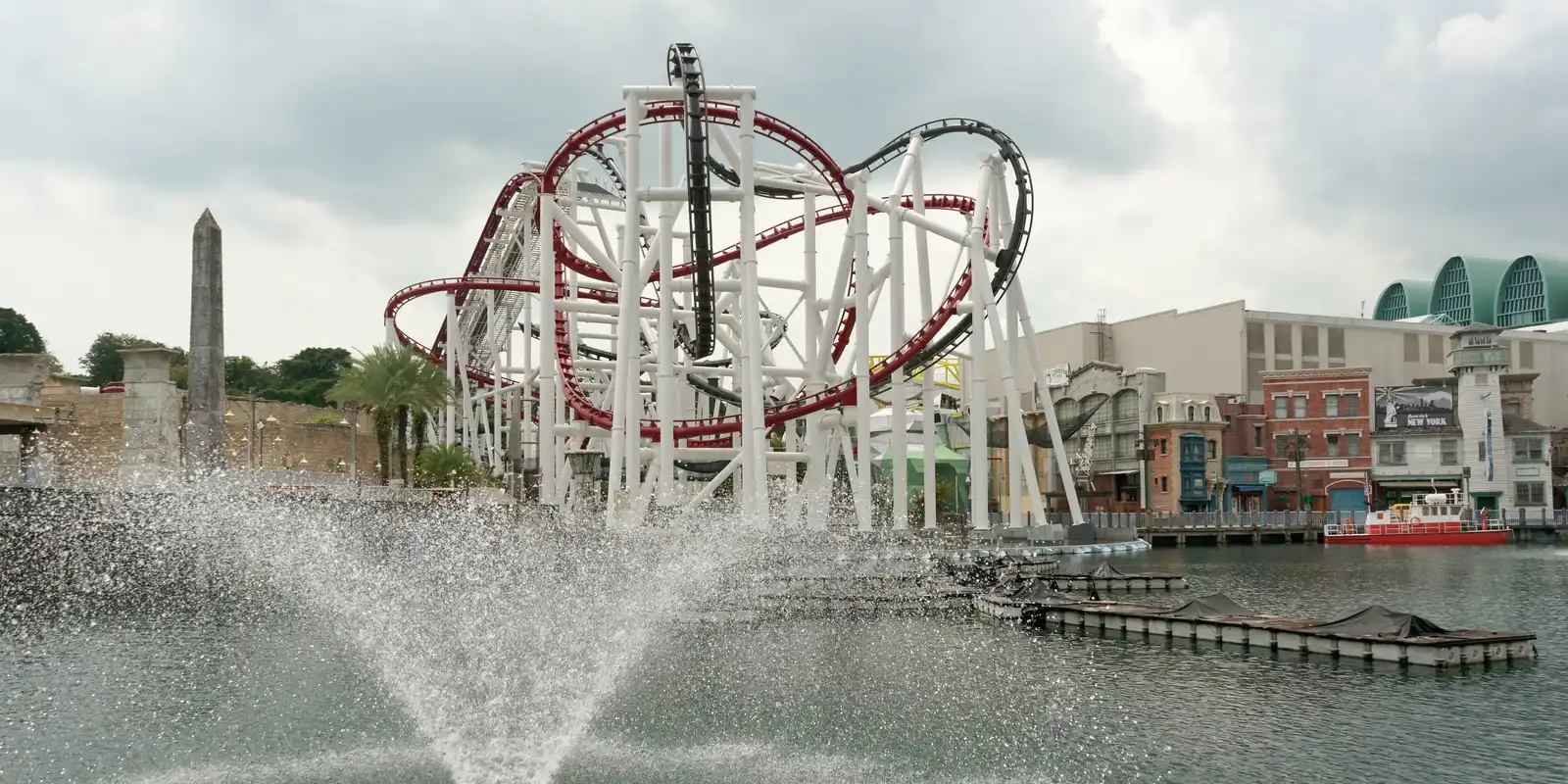 White steel supports of two intertwined roller coasters, one with red track and one with black track, rising behind a lagoon with a water fountain; a stone obelisk stands on the left and brick New York-style facades line the right bank