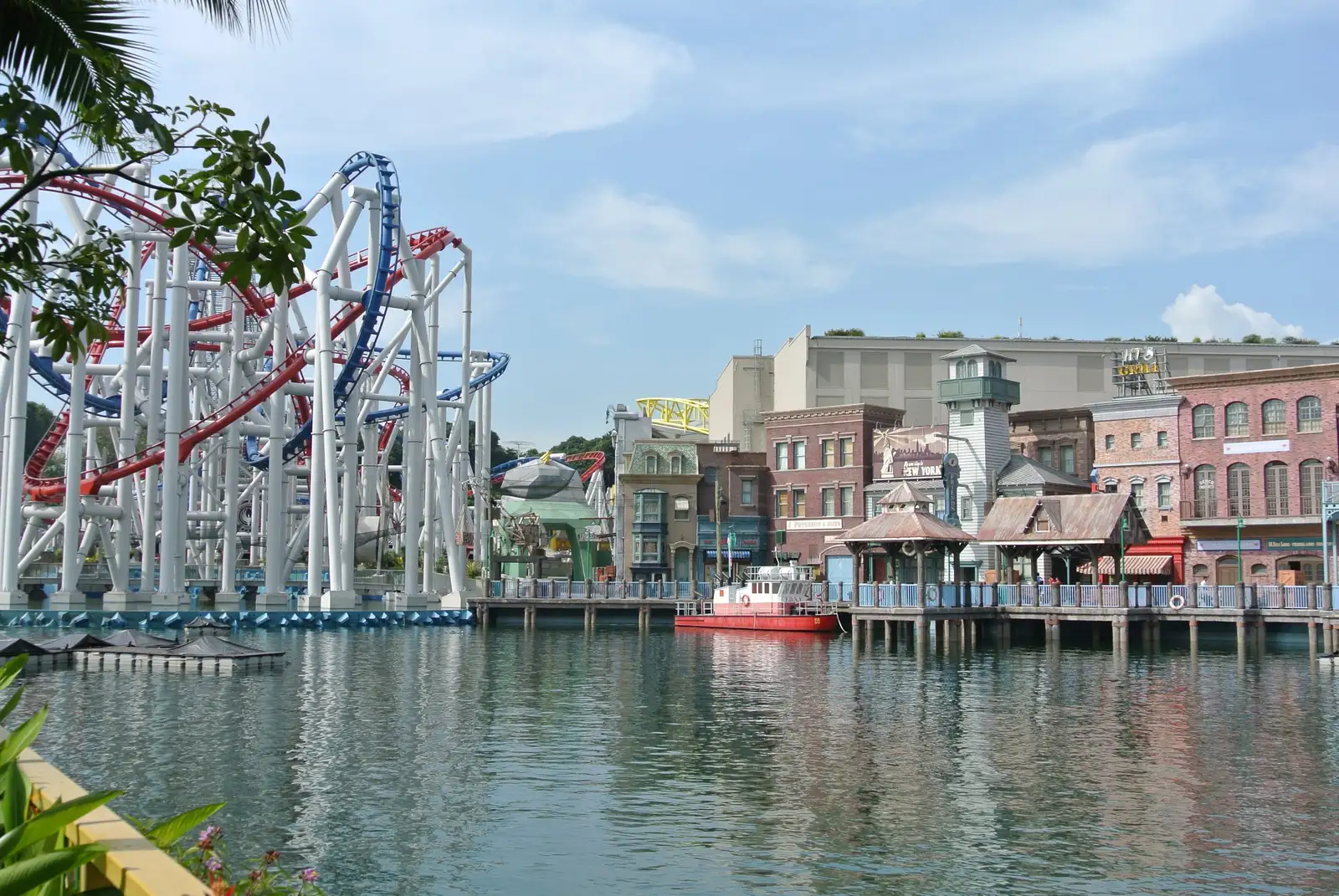 A calm lagoon reflecting a row of brick and clapboard New York-style buildings, with a small red fireboat moored at a wooden boardwalk and white roller-coaster supports on the left